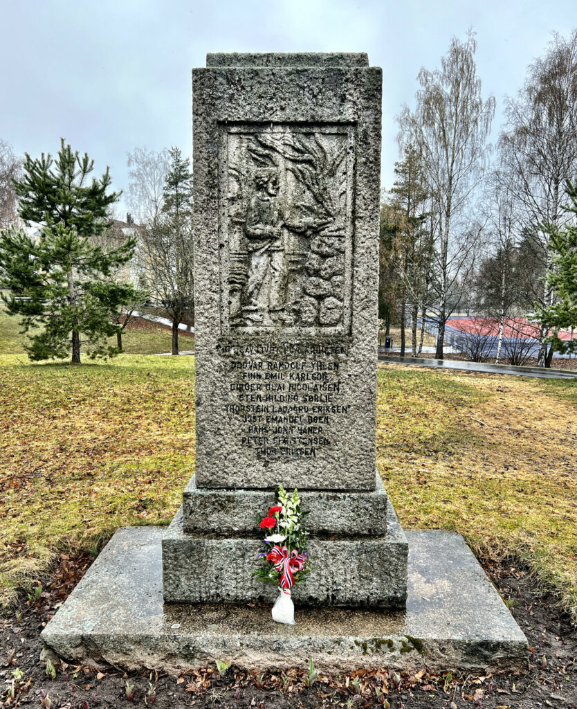 Bilde av bautaen over falne krigshelter som står på gressbakken vis-á-vis kirken på Høybråten. En blomsterbukett i friske farger er plassert på midten foran minnesmerket.