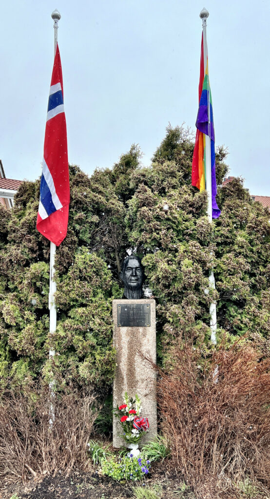 Bilde av minnesmerket over Karl Andersen foran Folkvang med det norske flagg og regnbueflagget på hver sin flaggstang på hver sin side av bysten. En blomsterbukett i friske farger er plassert i midten foran minnesmerket.